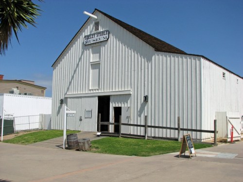 White wooden barn with a sign reading "Steely Stable Museum," surrounded by grass and a clear blue sky.