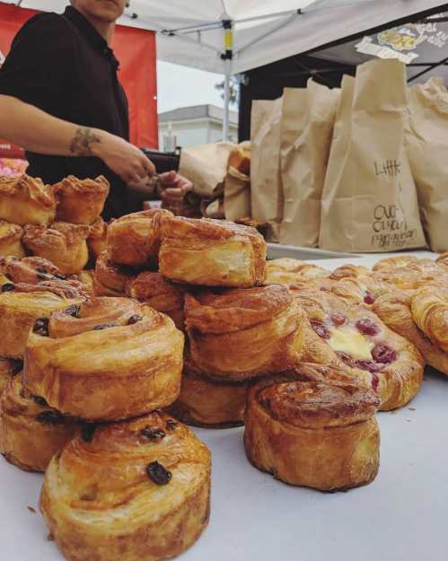 A variety of freshly baked pastries stacked on a table at a market, with a vendor in the background.