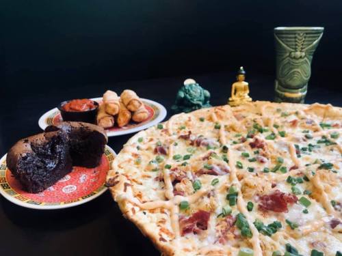 A pizza topped with green onions, a chocolate muffin, spring rolls, and a decorative green drink with statues in the background.