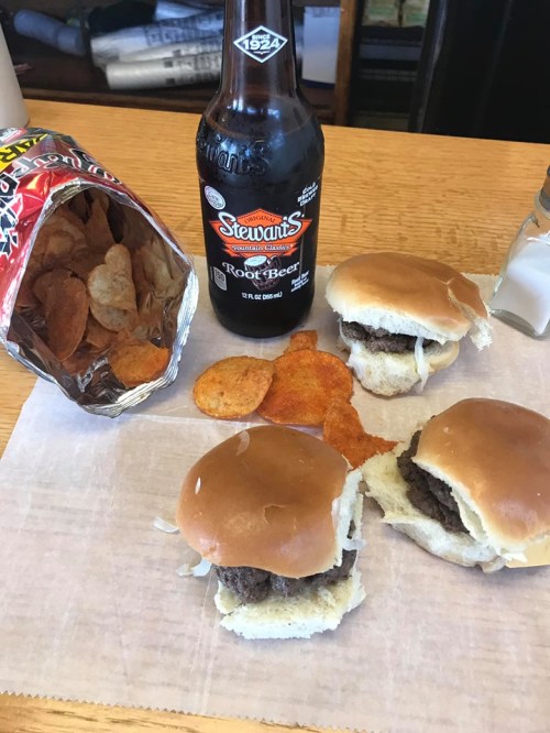 Three small burgers on a paper surface, a bag of chips, and a bottle of root beer.