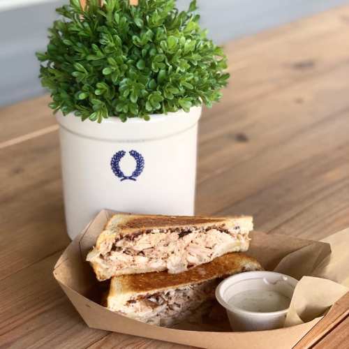 A toasted sandwich filled with chicken, served with a small cup of sauce, next to a potted plant on a wooden table.