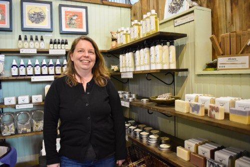 A smiling woman stands in a shop filled with natural products, including bottles, soaps, and candles on display.