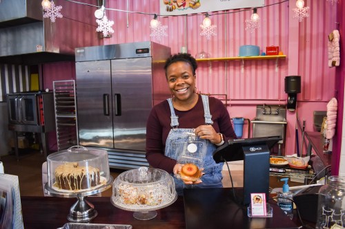 A smiling woman in overalls stands behind a counter with cakes and pastries, holding a donut in a colorful bakery.