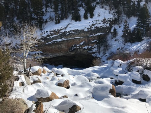 A snowy landscape with a river flowing through large rocks, leading to a cave entrance surrounded by trees.