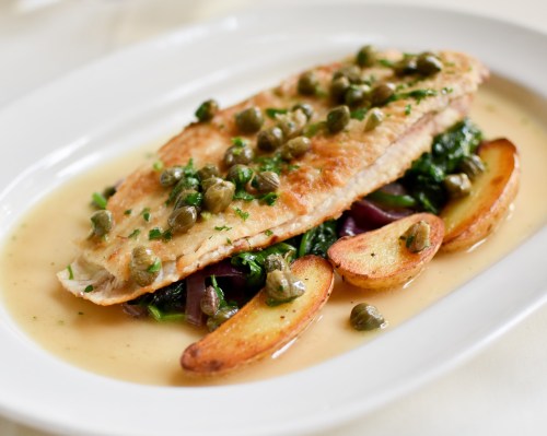A plated dish featuring a fillet of fish with capers, sautéed greens, and roasted potato wedges in a light sauce.