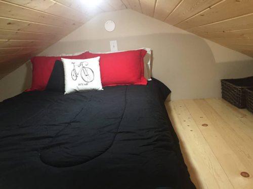 Cozy attic bedroom with a black bedspread, red pillows, and a decorative bicycle pillow against a sloped wooden ceiling.