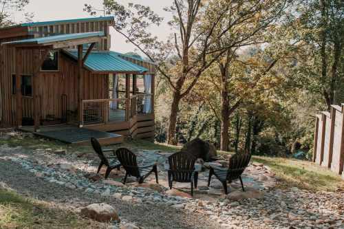 A cozy wooden cabin with a blue roof, surrounded by trees and a stone fire pit with chairs in the foreground.