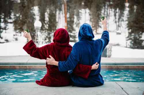 A couple in red and blue robes sit by a pool, raising glasses, with snowy trees in the background.