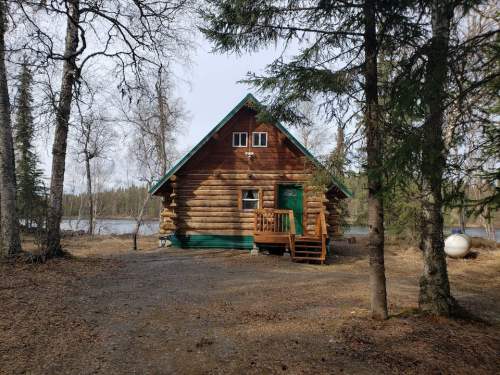 A rustic log cabin surrounded by trees, with a small porch and a view of a river in the background.