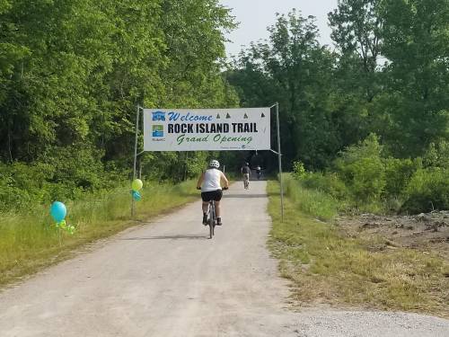 A cyclist rides under a banner welcoming visitors to the Rock Island Trail's grand opening, surrounded by greenery.