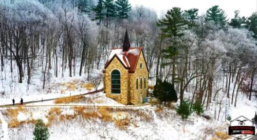 A small stone church with a steeple surrounded by snow-covered trees and a path, with two people walking nearby.