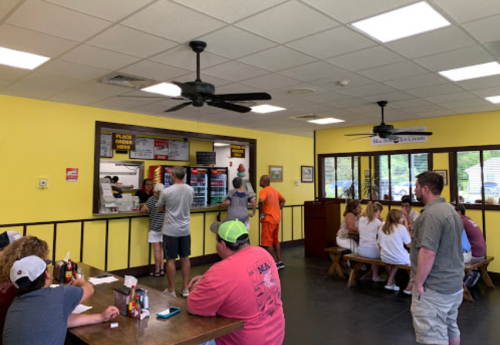 A busy ice cream shop with customers ordering at the counter and others seated at tables. Bright yellow walls.