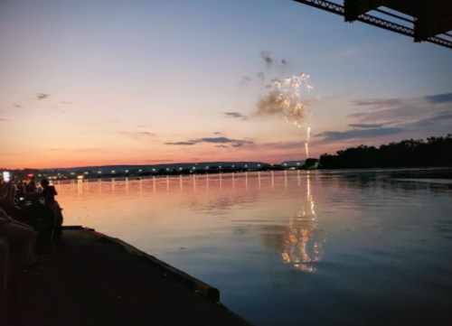 A sunset over a river with fireworks reflecting on the water, and silhouettes of people watching from the shore.