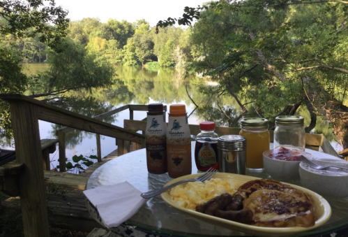 A serene breakfast scene by a lake, featuring eggs, sausage, French toast, and drinks on a glass table surrounded by nature.