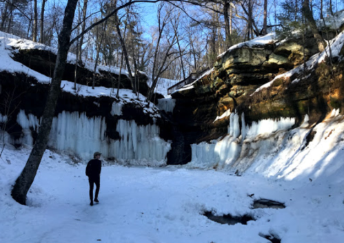 A person walks through a snowy landscape with icy cliffs and frozen waterfalls under a clear blue sky.