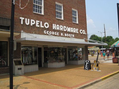 Exterior of Tupelo Hardware Co. with a sign reading "OPEN" and a guitar display outside. Sunny day, people in background.