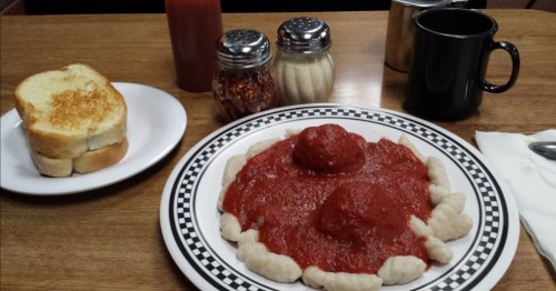 A plate of gnocchi topped with marinara sauce and meatballs, served with toast and condiments on a wooden table.