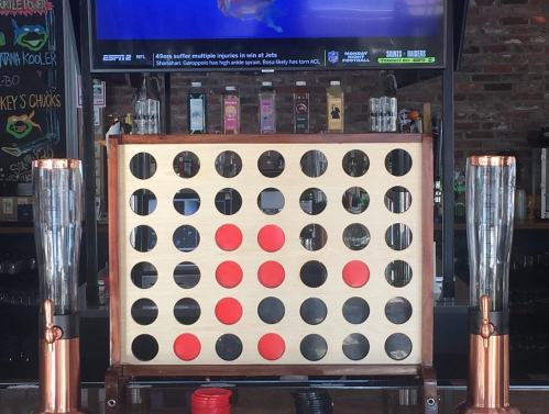 A large Connect Four game set up in a bar, with red and black discs ready for play. Behind it, a TV displays sports.