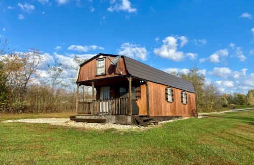 A small wooden cabin with a porch, set against a blue sky and grassy landscape. Trees are visible in the background.