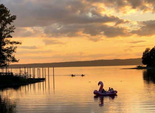 A pink swan float and kayakers on a calm lake at sunset, with colorful clouds reflecting on the water.