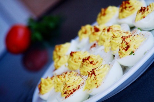 A platter of deviled eggs topped with yellow filling and paprika, with a tomato and herbs in the background.