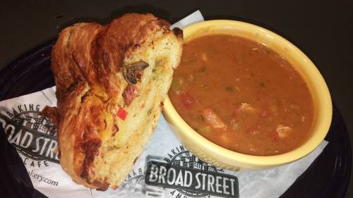 A bowl of hearty soup with vegetables and meat, served alongside a flaky, savory pastry on a branded paper.