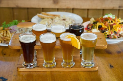 A flight of seven craft beers on a wooden paddle, with a plate of food in the background.