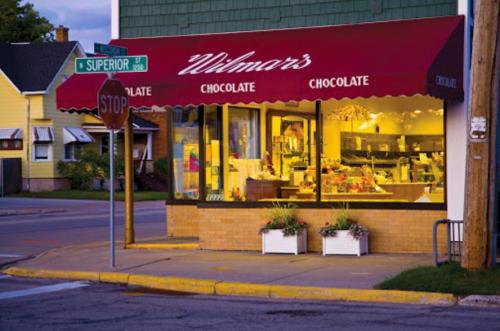 A cozy chocolate shop with a red awning, warmly lit windows, and flower planters at the corner of a street.
