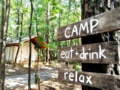 A wooden signpost in a forest, directing to "CAMP," "eat + drink," and "relax," with a cabin in the background.