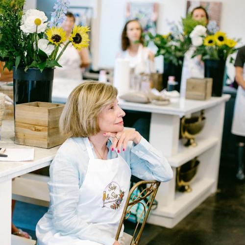 A woman in an apron sits thoughtfully in a flower arranging class, surrounded by participants and floral arrangements.