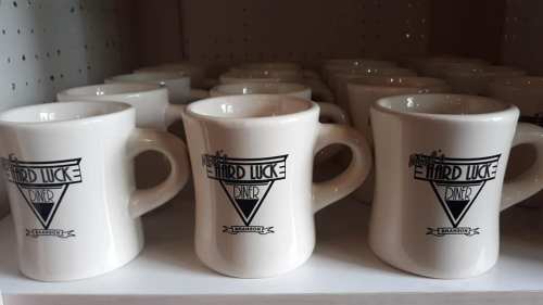 A row of white coffee mugs with the logo of "Hard Luck Diner" displayed on a shelf.