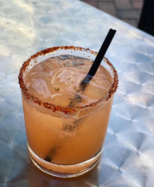 A refreshing cocktail in a glass with a spicy rim, filled with ice and a black straw, on a metallic surface.
