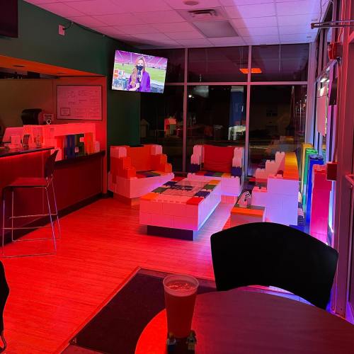 A colorful lounge area with block-style furniture, a TV in the background, and ambient lighting. A drink sits on a table.