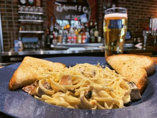 A plate of creamy pasta with grilled chicken, served with two slices of toasted bread and a glass of beer in the background.