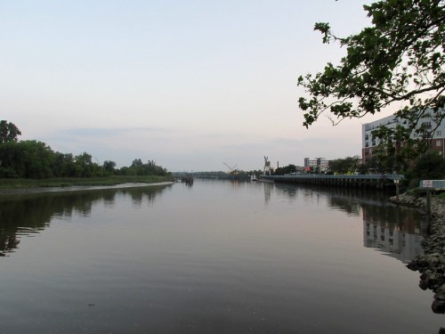 A calm river reflects the sky, bordered by trees and buildings, under a soft evening light.