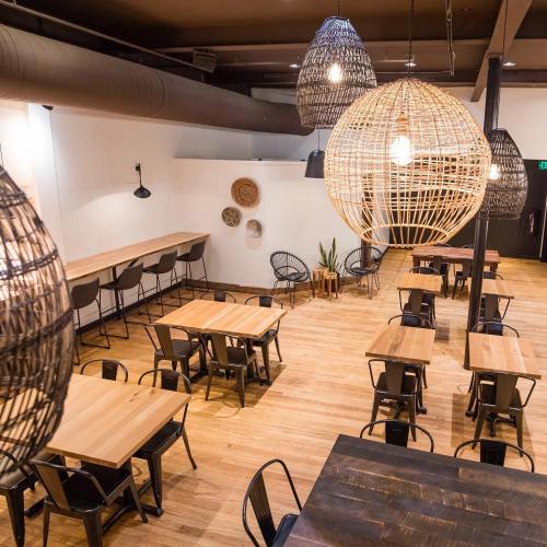 A modern restaurant interior with wooden tables, black chairs, and large woven pendant lights.