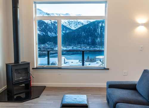 Cozy interior with a wood stove, a view of mountains and a lake through a large window, and a gray sofa.
