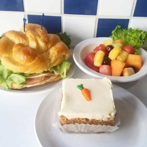 A croissant sandwich with lettuce and turkey, a bowl of mixed fruit, and a slice of carrot cake with a small carrot decoration.
