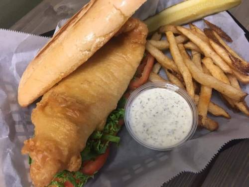 A crispy fish sandwich with lettuce and tomato, served with fries, a pickle, and a side of tartar sauce.