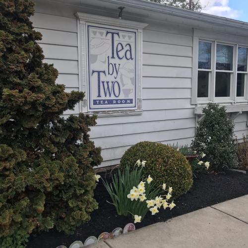 A quaint tea room named "Tea by Two" with a sign, surrounded by greenery and blooming flowers.