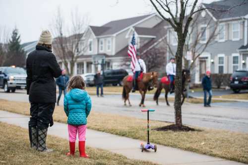 A child in a blue jacket watches a parade with horses and riders, while an adult stands nearby on a suburban street.