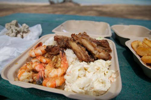 A takeout container filled with shrimp, grilled meat, rice, and macaroni salad on a green surface.