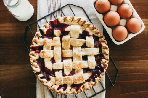 A freshly baked lattice-top pie on a cooling rack, with a carton of brown eggs and a bottle of milk nearby.