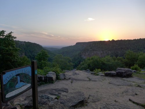 Sunset over a valley with a sign for Cedar Falls Trail in the foreground, surrounded by trees and rocky terrain.