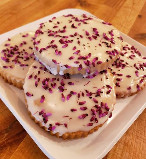Three round cookies with a glossy icing topped with pink sprinkles, arranged on a white plate on a wooden surface.