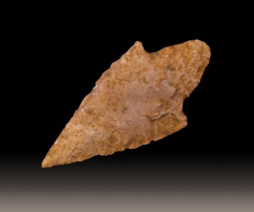A pointed stone artifact, likely an arrowhead, with a smooth surface and natural texture, set against a dark background.