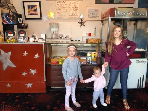 Two girls stand in a vintage-style shop, holding hands, with a display of snacks and a retro popcorn machine behind them.