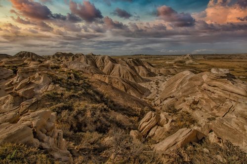 A rugged landscape with layered rock formations under a colorful sky at sunset, featuring scattered vegetation.