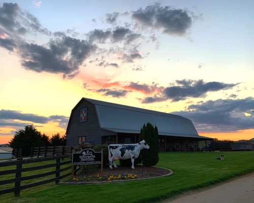 A barn with a cow statue in front, set against a colorful sunset and cloudy sky. Green grass and flowers in the foreground.