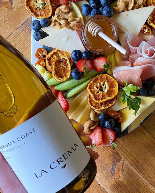 A hand holds a bottle of La Crema Chardonnay next to a colorful cheese and fruit platter with nuts and honey.